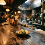 Otoshi: The Food You Didn't Order A narrow izakaya counter at night. A small dish sits in front of an empty seat. The owner's hands are already moving on to the next task.