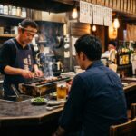 Hitori Nomi: The Japanese Art of Drinking Alone A single man at an izakaya counter, seen from behind. He sits upright, a glass of beer in front of him, facing the owner who is working quietly. The counter is half-empty. Warm light. Late evening.