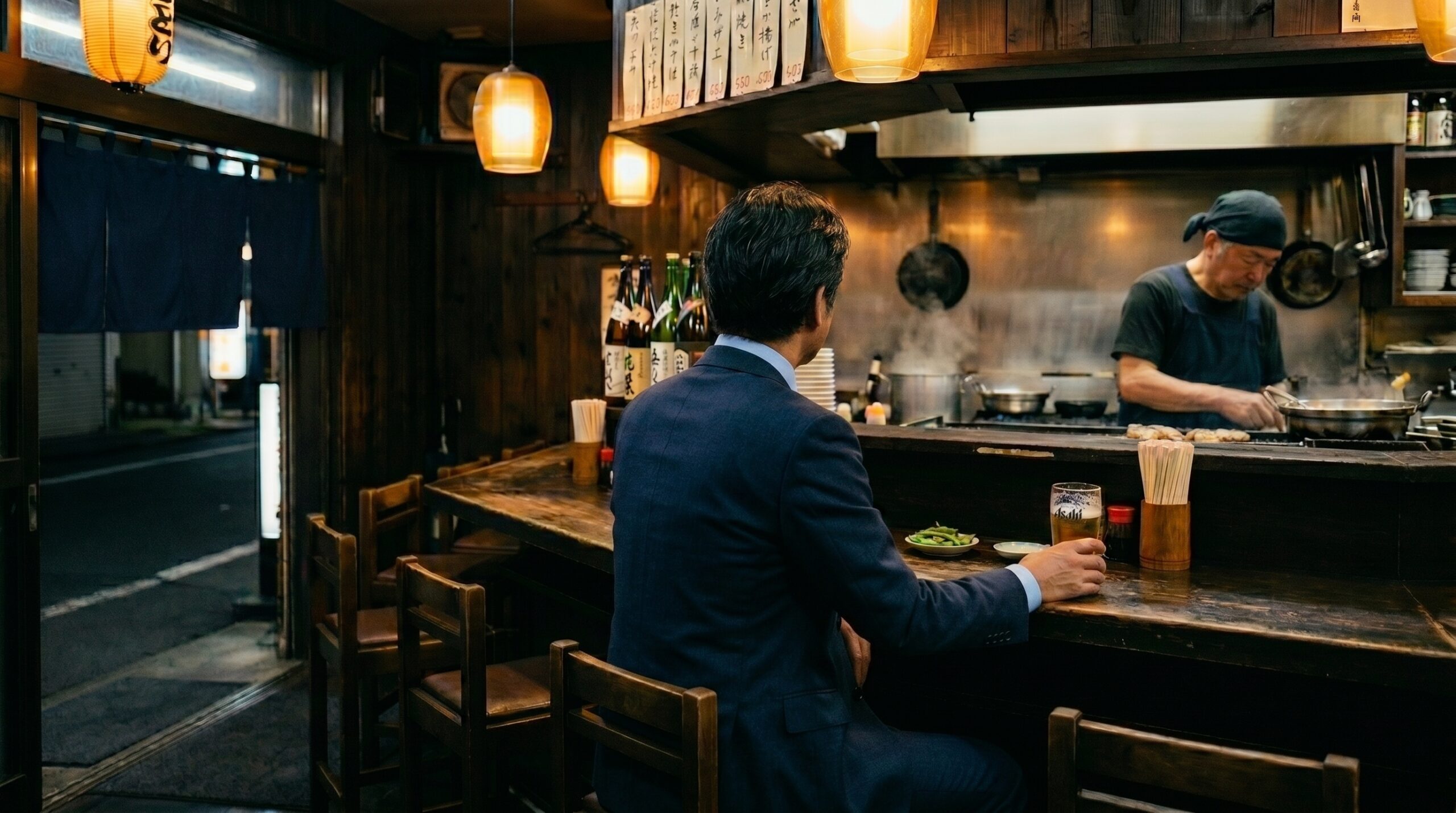 A man sits alone at a four-seat izakaya counter, late evening. A glass of beer in front of him, the owner working quietly in the background. He looks completely at ease.