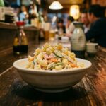 Macaroni Salad: The Dish That Reveals a Kitchen A small dish of Japanese-style macaroni salad in a plain white ceramic bowl on a dark izakaya counter. Simple, unpretentious. A toothpick rests on the rim. Warm ambient light.