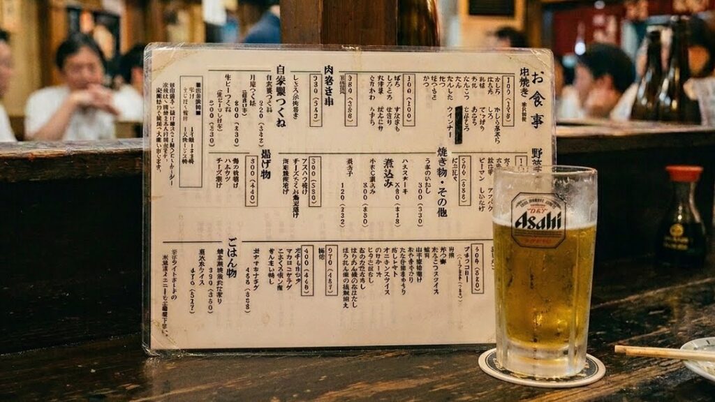 A simple Japanese izakaya menu — a single laminated sheet with handwritten kanji and small printed text. No photos. A glass of beer beside it. The counter is dark.