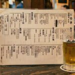 A simple Japanese izakaya menu — a single laminated sheet with handwritten kanji and small printed text. No photos. A glass of beer beside it. The counter is dark.