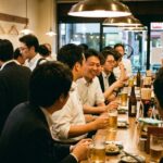 A Japanese standing bar — tachinomi — at early evening. People standing at a narrow counter along the wall, drinks in hand. The space is small, efficient, busy. Nobody is sitting. Nobody seems to mind.