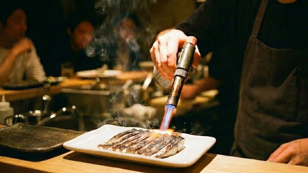 A torch being applied to shimesaba skin at an izakaya counter — the small flame catching the skin, blistering it in real time. The fish is on a plate below. The cook watches closely.