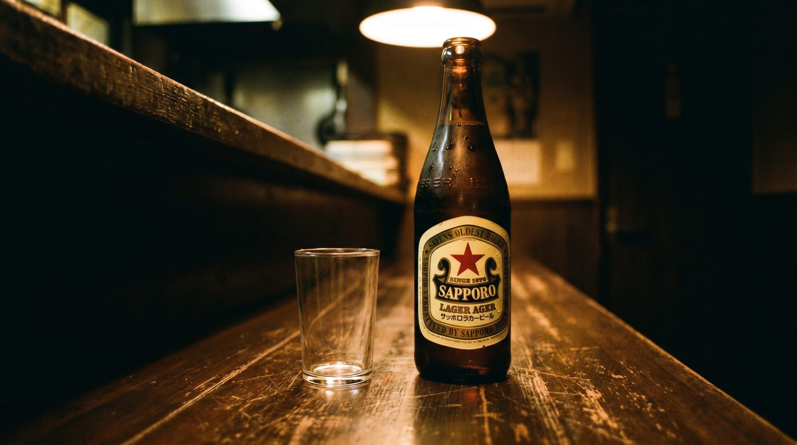 A classic Sapporo Lager bottle — the red star label — on a worn izakaya counter. Dark wood, warm light, nothing decorative. The bottle is cold. The glass beside it is plain.