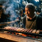 Yakitori and Yakiton: Two Different Things at the Same Counter A charcoal grill behind an izakaya counter — skewers lined up over the coals, some chicken, some pork offal. The smoke rises. The owner stands behind it, watching. The light is from the coals.