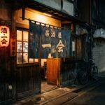 The entrance to a small Japanese izakaya at night. 暖簾 (noren curtain) hangs in the doorway, slightly worn. Warm light glows from inside. The street outside is empty. The feeling of a place that knows who belongs and who doesn't.