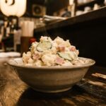 A generous serving of izakaya potato salad in a small bowl — creamy, slightly chunky, with visible cucumber and ham. A simple bowl on a worn counter. No garnish. Just the thing itself.