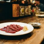 A plate of basashi — thin slices of raw horsemeat, deep red, arranged simply on a white plate. A small mound of grated ginger beside it. A dish of soy sauce. Nothing decorative.