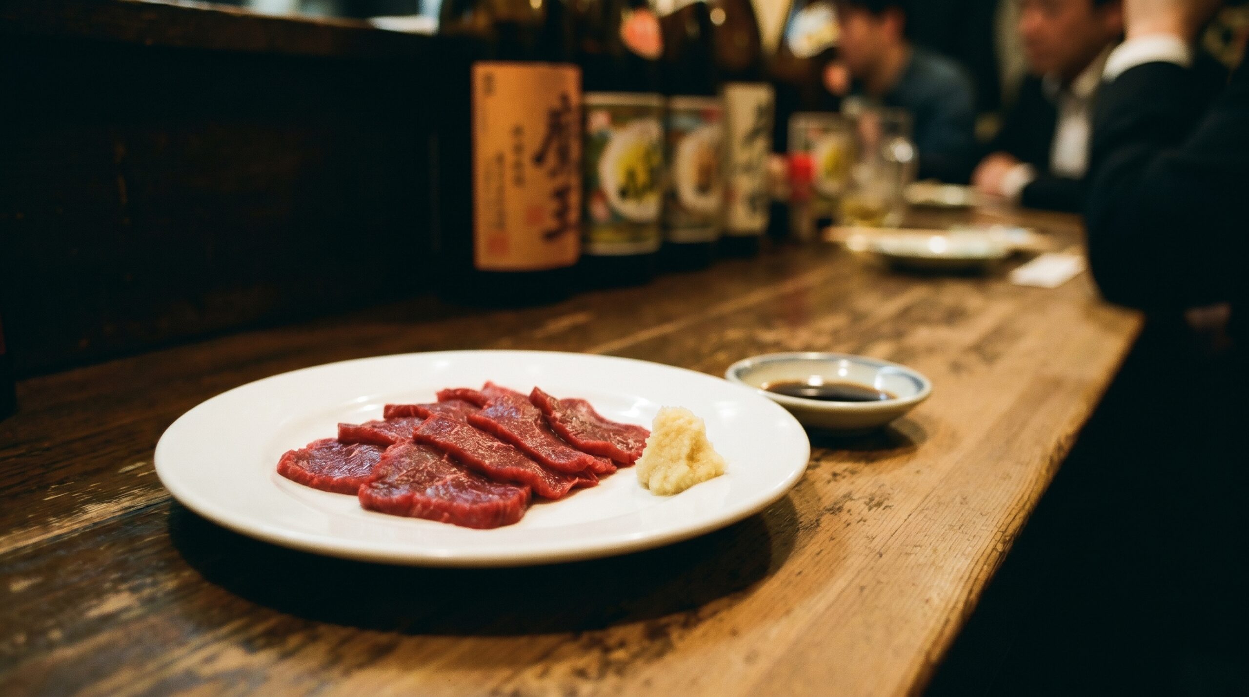 Basashi: Raw Horsemeat, and Why a Place That Does It Right Doesn't Explain It 1 A plate of basashi — thin slices of raw horsemeat, deep red, arranged simply on a white plate. A small mound of grated ginger beside it. A dish of soy sauce. Nothing decorative.
