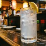 Lemon Sour: Japan's Default Drink, and What That Means A tall glass of lemon sour on a dark izakaya counter — condensation on the glass, a wedge of real lemon on the rim, ice visible inside. The drink is pale gold. The light is warm.