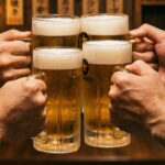 Four glasses of beer raised mid-kanpai around an izakaya table. The glasses are touching at the tops. Faces are slightly blurred. The moment just before everyone drinks. Warm light.
