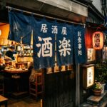 Noren: What Happens When You Push Through the Curtain A dark blue noren curtain hanging in the entrance of a small izakaya at night. The curtain is slightly parted, warm light visible inside. The street outside is quiet. The curtain is worn at the edges.