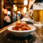 A small dish of chanja — deep orange-red, glistening, fermented cod entrails — in a plain white ceramic dish on a dark izakaya counter. A toothpick beside it. A cold beer glass in the background.