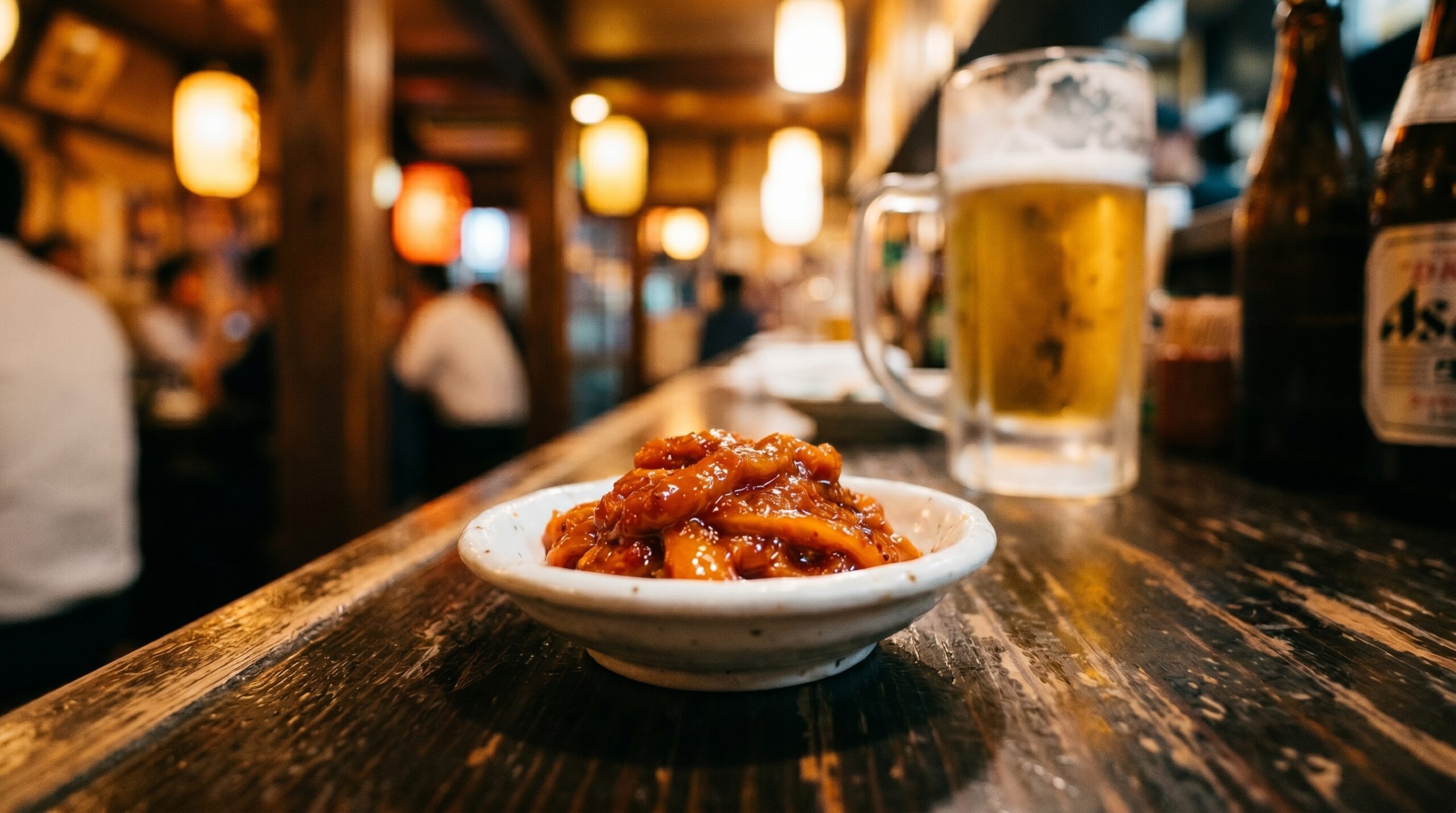A small dish of chanja — deep orange-red, glistening, fermented cod entrails — in a plain white ceramic dish on a dark izakaya counter. A toothpick beside it. A cold beer glass in the background.