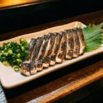 lices of aburi shimesaba on a small plate — the skin slightly charred and blistered from the torch, the flesh below still silvery and cured. A small mound of grated ginger beside it. Clean, precise presentation.